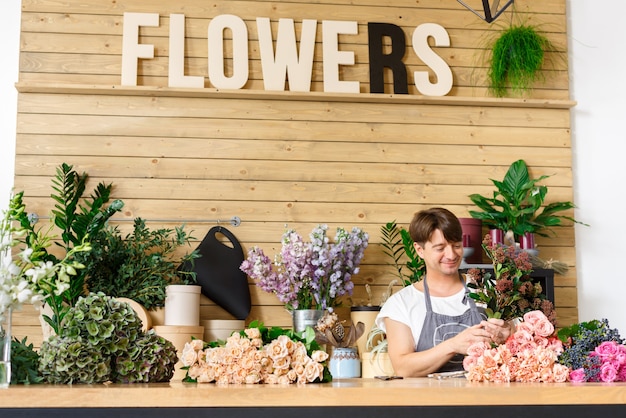 写真 小規模なビジネス。フラワーショップのカウンターデスクでバラの花束を作る男性の花屋。花のデザインスタジオの男性アシスタントまたはオーナー。装飾やアレンジを行っています。花の配達、注文の作成