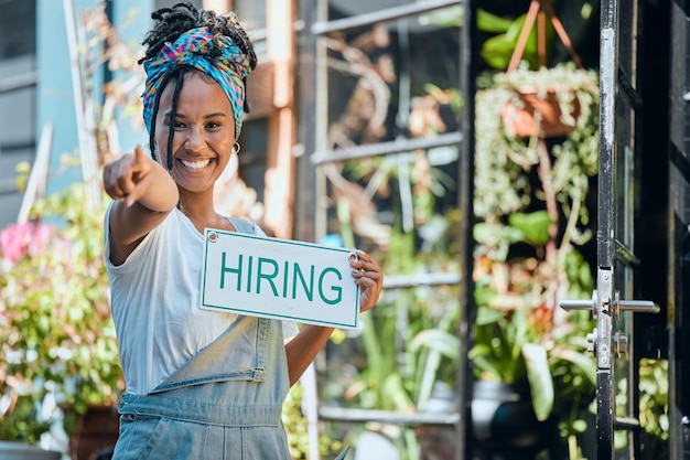 Photo small business manager or black woman with a hiring sign at door entrance in floral retail store advertising marketing and happy entrepreneur smiles pointing with job offer message at flower shop