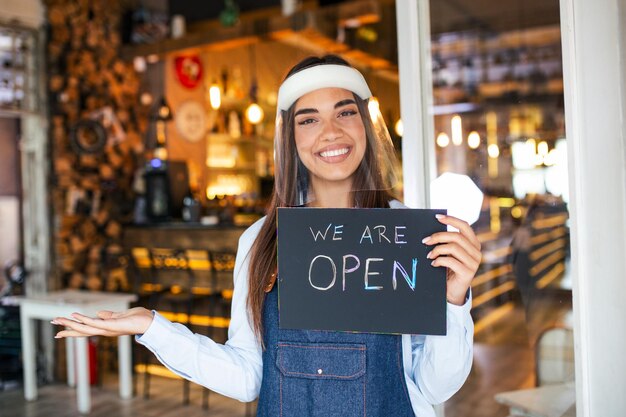 Photo small business owner smiling while holding the sign for the reopening of the place after the quarantine due to covid19 woman with face shield holding sign we are open support local business