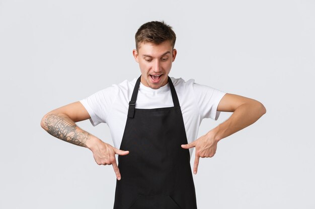 Photo small business owners, coffee shop and staff concept. enthusiastic happy and surprised handsome cafe barista, waiter in black apron pointing fingers down and looking with temptation bottom banner