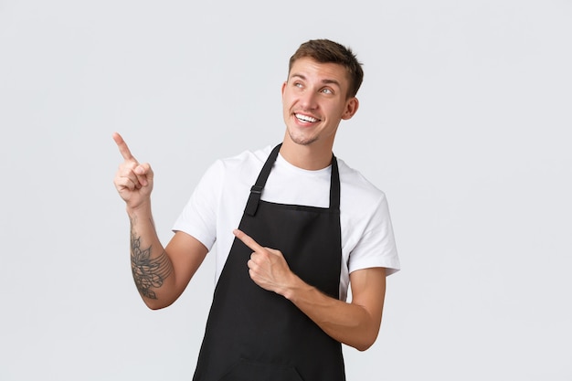 Small business owners, coffee shop and staff concept. Smiling pleased and happy handsome barista in black apron, cafe employee pointing and looking upper left corner satisfied, showing advertisement