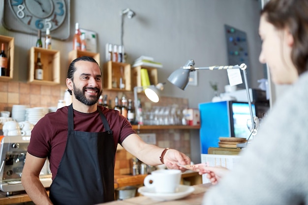 写真 中小企業、人々、サービスのコンセプト – バーで顧客にお金を与えるコーヒーカップを持つ幸せな男性またはウェイター