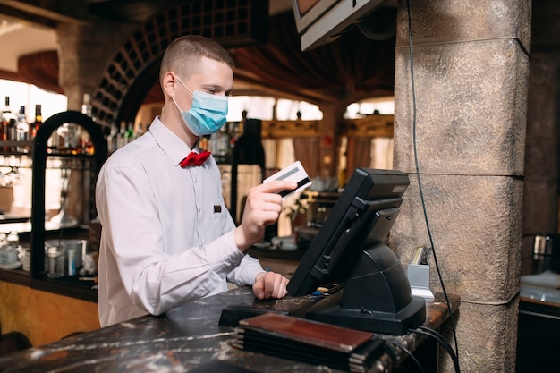 Small business, people and service concept. man or waiter in medical mask at counter with cashbox working at bar or coffee shop.