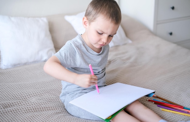 A small caucasian boy in a gray tshirt and shorts on the bed draws with colored pencils in a
