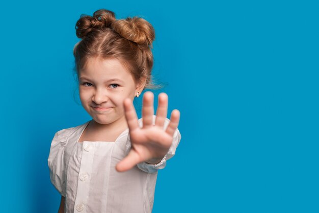 Small caucasian girl is gesturing the stop sign and smile at camera while posing on a blue studio wall with free space