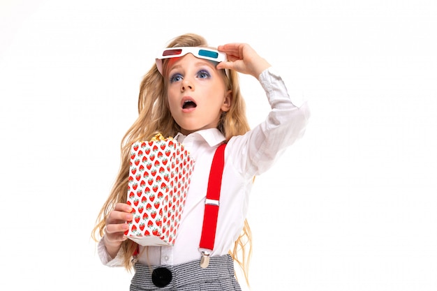 Small caucasian girl with makeup and long blonde hair with pop-corn and 3D glasses is shocked