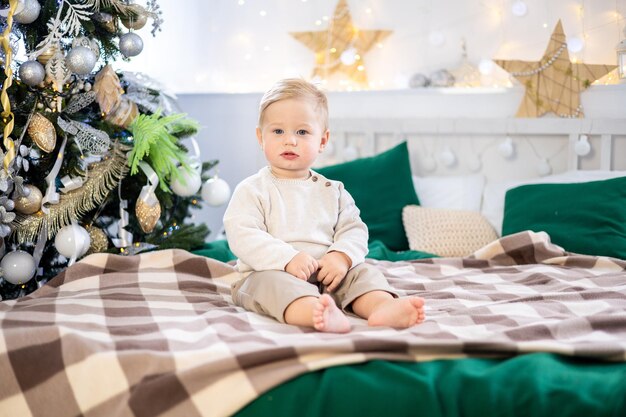 니트 스웨터를 입은 어린 소년이 집에서 축제로 장식된 크리스마스 트리를 배경으로 침대에 앉아 있고 그 아이는 집에서 크리스마스와 새해를 축하하고 있습니다