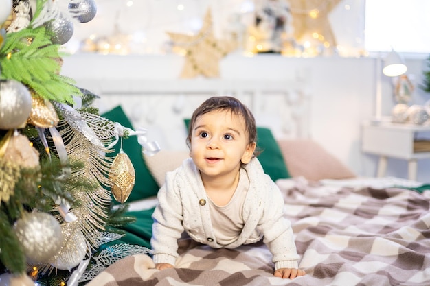 A small child boy in a knitted sweater is sitting on the bed against the background of a festive decorated Christmas tree at home the kid is celebrating Christmas and New year at home