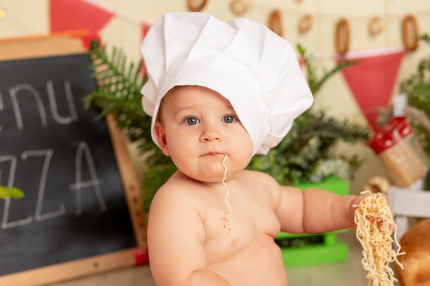 A small child cook in a hat in the kitchen among the products and eating spaghetti with his hands