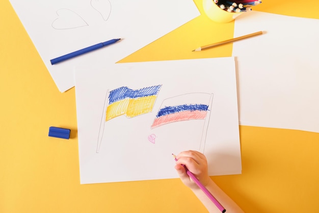 A small child draws a heart on a sheet the flags of russia and ukraine a peaceful solution to the military conflict between countries concept yellow background faith in a better future