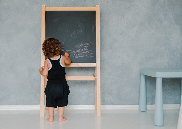 Маленький ребенок рисует мелом на черном меловой доске дома в питомнике на фоне серой стены.