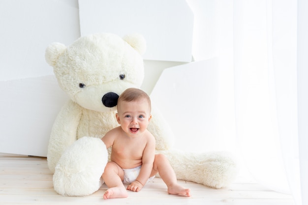 Photo a small child a girl of 6 months is sitting with a large soft bear in a bright apartment in diapers