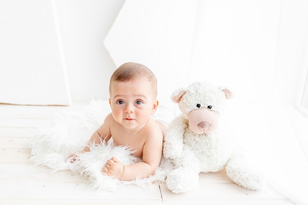 A small child a girl of 6 months is sitting with a large soft bear in a bright apartment in diapers