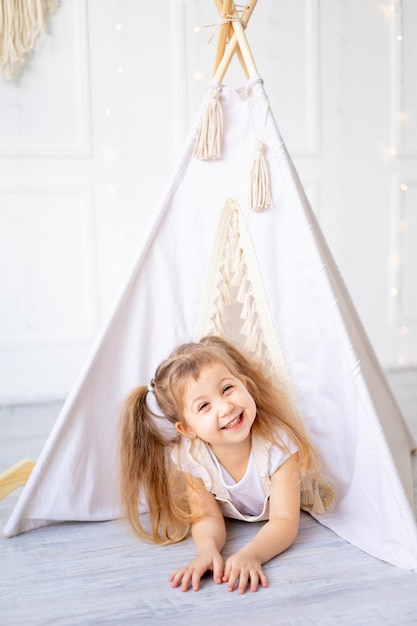 A small child girl looks out of the wigwam at home in a bright children's room and laughs