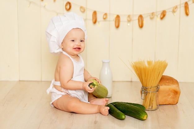 Un bambino piccolo, una bambina di sei mesi, è seduto in cucina con indosso un cappello da cuoco e un grembiule e mangia una zucca verde