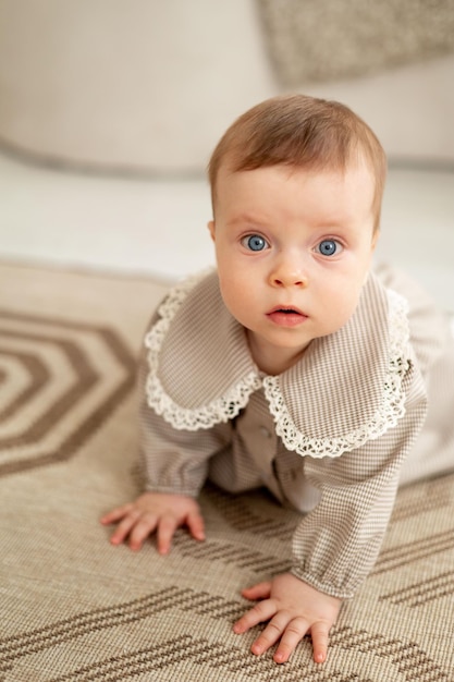 Маленькая девочка с голубыми глазами в бежевом платье ползает по ковру дома.