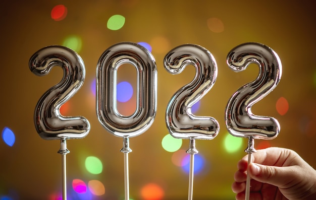 Small child hand is holding the last symbol 2 in the number 2022 on a dark golden background with a bokeh. Concept of Happy New Year greetings