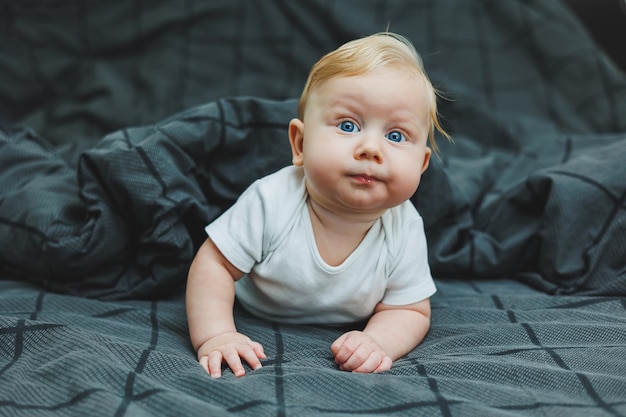 작은 아이가 집에서 큰 침대 위에 누워 있습니다. 회색 침대에 누워있는 5 개월 된 아기의 초상화 즐거운 행복한 아이