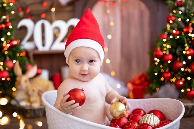 A small child is sitting in a bath with Christmas tree toys with Christmas trees. A little girl in a red Santa Claus hat. Happy Holidays, New Year. Christmas Time