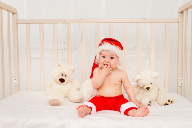 A small child is sitting in a crib in a Santa hat with stuffed toys bears