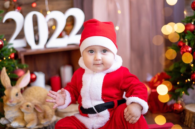 A small child is sitting on a sled under the Christmas trees. A large portrait. A baby in a red Santa Claus costume with a gift in a gift box. Happy Holidays, New Year. Christmas Time