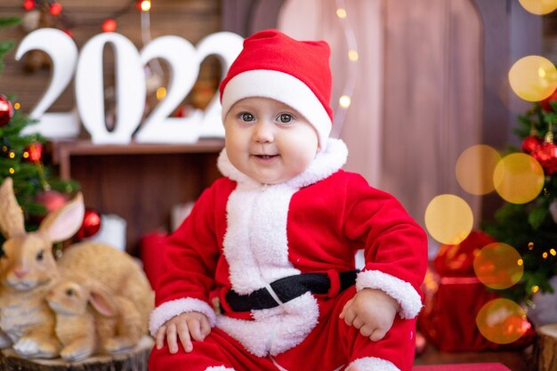 A small child is sitting on a sled under the Christmas trees. A large portrait. A baby in a red Santa Claus costume with a gift in a gift box. Happy Holidays, New Year. Christmas Time