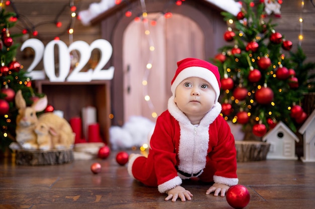 A small child is sitting on a sled under the Christmas trees. A large portrait. A baby in a red Santa Claus costume with a gift in a gift box. Happy Holidays, New Year. Christmas Time