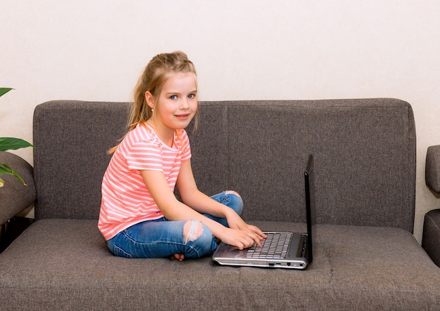 A small child is surfing the Internet while sitting on a gray sofa. girl with laptop on the couch sitting