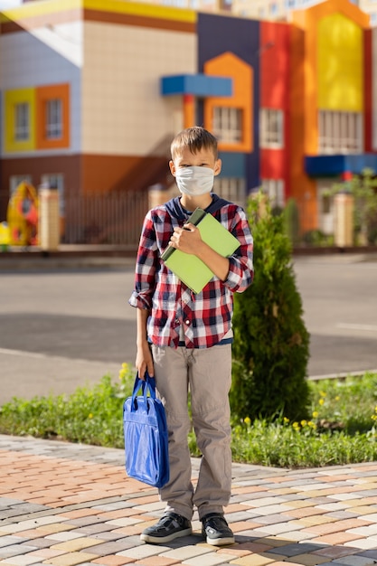 A small child in a mask with a school bag during an outbreak of the virus near the school wall