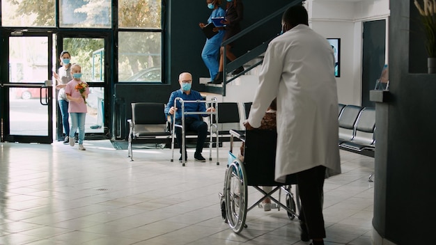 Foto bambino piccolo e madre che visitano un paziente anziano presso la struttura, portando fiori alla donna in sedia a rotelle. bambino che viene a vedere un adulto anziano con disabilità nel centro sanitario medico, coronavirus.