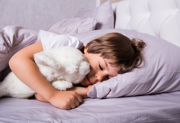 A small child in pajamas sleeps under a blanket on a bed with a white stuffed toy