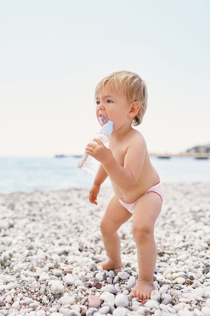 Un bambino piccolo in pantaloncini sta su una spiaggia di ciottoli con una bottiglia d'acqua in mano Foto Premium