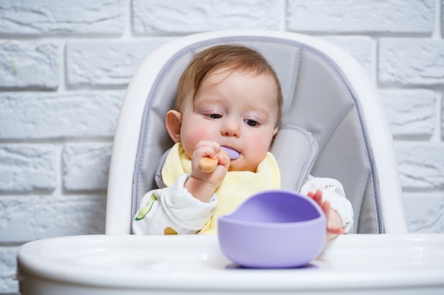 小さな子供がハイチェアに座って、スプーンで皿から食べ物を食べます。赤ちゃんを養うための赤ちゃん用シリコーン器具