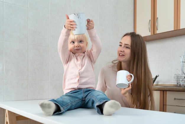 Small child in surprise looks at mug sitting next to mom in kitchen