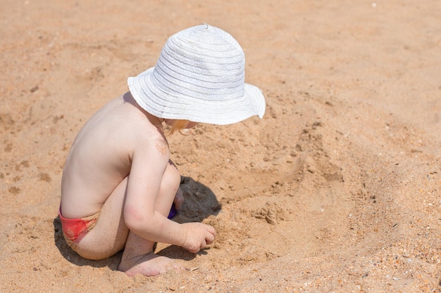 白いパナマと水泳パンツを着た小さな子供が、休日に海岸の砂で遊んでいます。