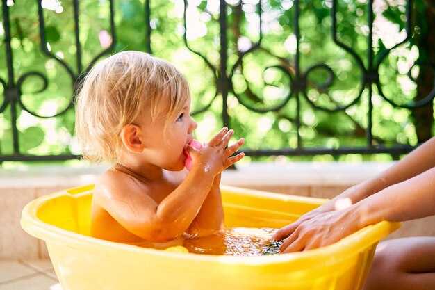 Un bambino piccolo con un giocattolo in mano siede in una bacinella d'acqua sul balcone Foto Premium