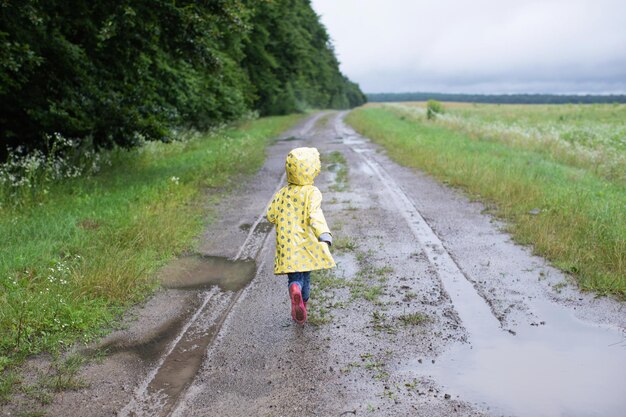 黄色いレインコートを着た小さな子供が未舗装の道路と泥の上を走る