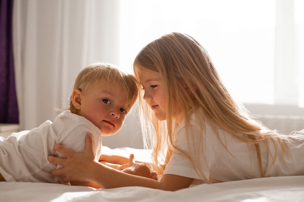 small children brother and sister in white Tshirts are lying on the bed and looking at each other