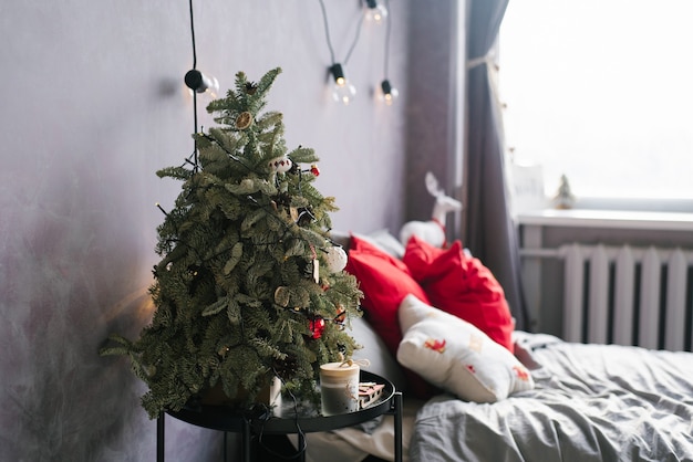 Маленькая новогодняя елка на тумбочке и подарки возле кровати в стильной спальне