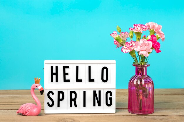 Piccoli garofani rosa colorati in vaso e scatola luminosa con testo ciao primavera, figura di fenicottero sul tavolo di legno bianco e parete blu holiday card concetto stagionale
