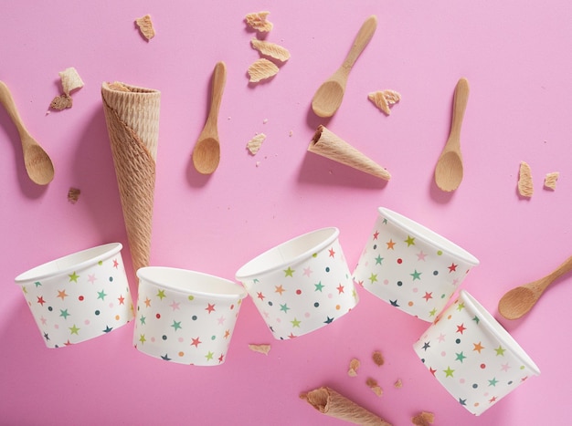 Small cups and ice cream cones empty and wooden spoons on plain pink background top view