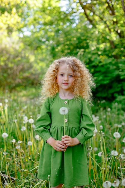 A small curly haired blonde girl blows on dandelions in a clearing