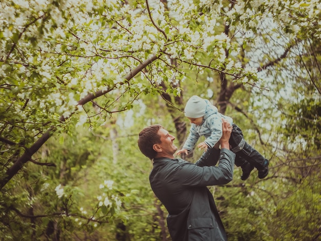봄 야외 공원에서 산책하는 그의 아버지와 함께 작은 귀여운 아기. 남자는 그의 손에 그의 작은 아들을 올립니다.