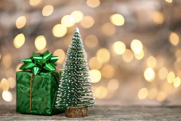 Small decorative Christmas trees and gift box on wooden table against blurred lights