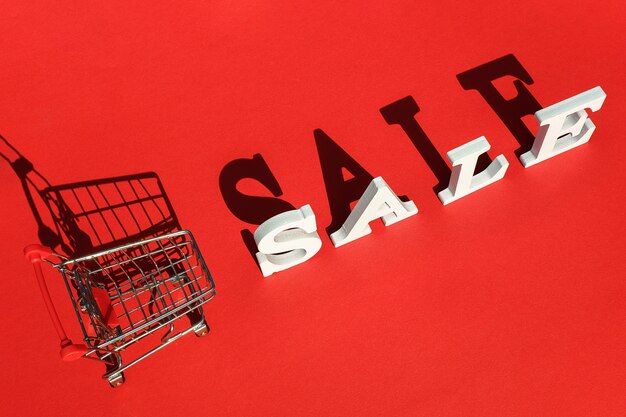 Photo small empty shopping trolley cart and word sale of white letters casts a large shadow on red background