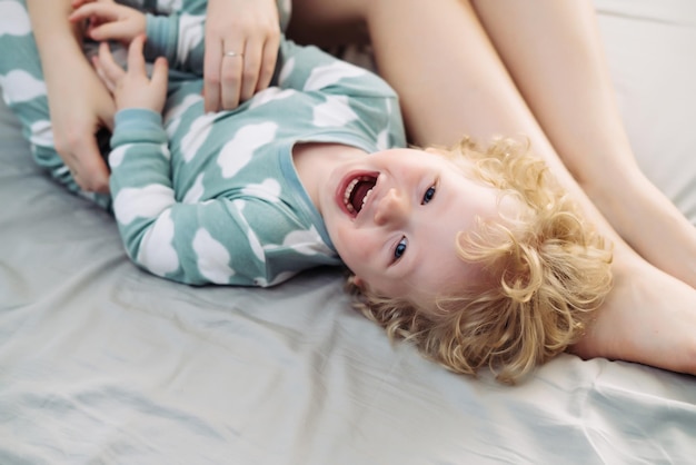 Small fairhaired child boy in blue pajamas laughing lying on bed next to mom