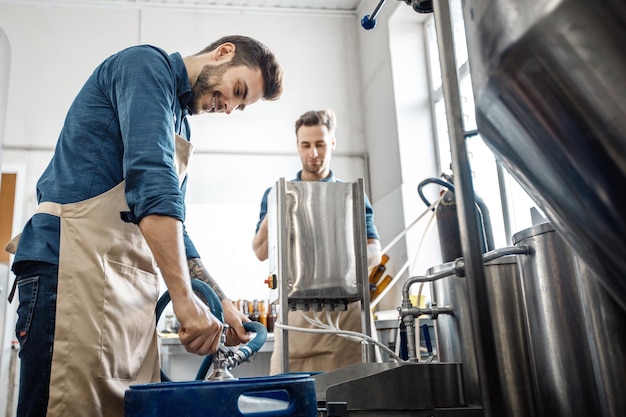 Small family business, drink productions and modern brewery. Busy young attractive male workers in aprons work with beer brewing equipment and making good lager in factory interior, free space