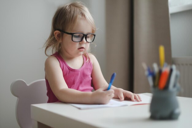 テーブルで書いたり描いたりする眼鏡の小さな面白い女の子