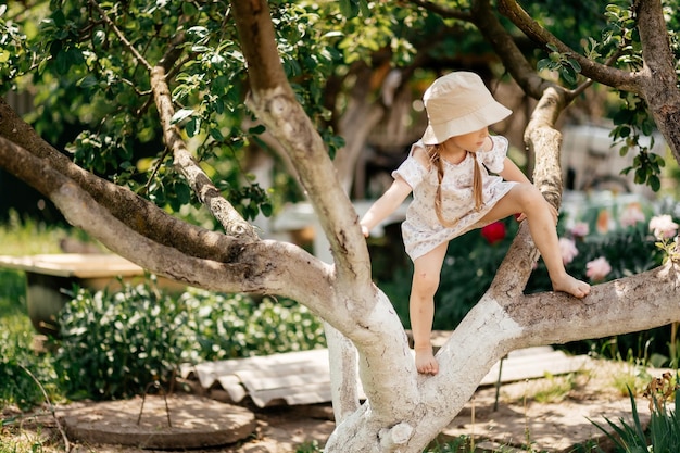 Small girl climb tree in summer garden activity Little girl on tree branch childhood Vacation activity lifestyle Kid fashion style beauty Childhood youth growth
