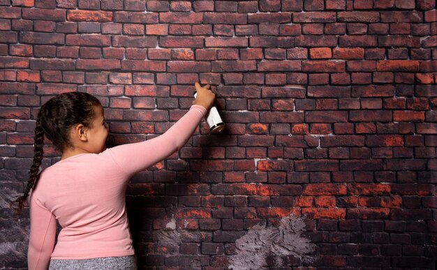 Foto piccola ragazza che fa pose con uno spray davanti a un vecchio sfondo di un muro di mattoni, sfondo scuro, messa a fuoco selettiva.